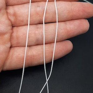 May include: Three white elastic cords, approximately 45 cm long, are held in a hand against a black background.