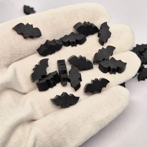 May include: A collection of small, black bat-shaped embellishments. The bats are scattered on a white surface, with some appearing to be held in a gloved hand. The bats have a simple, stylized design, perfect for Halloween or craft projects.