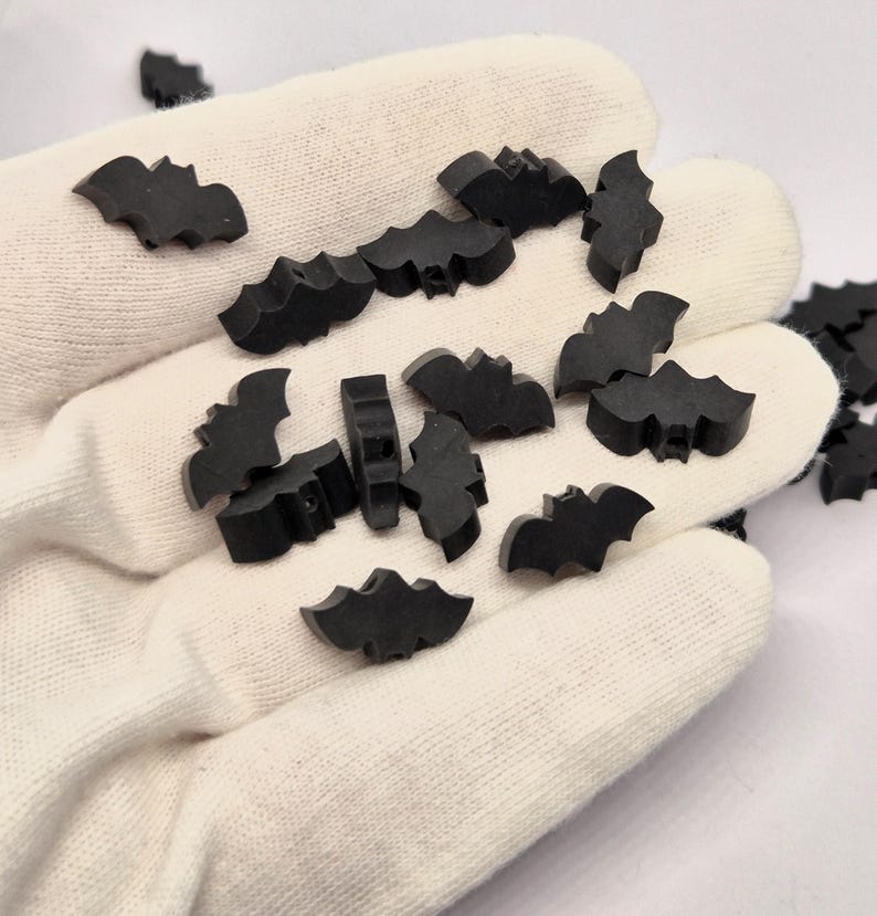 May include: A collection of small, black bat-shaped embellishments. The bats are scattered on a white surface, with some appearing to be held in a gloved hand. The bats have a simple, stylized design, perfect for Halloween or craft projects.