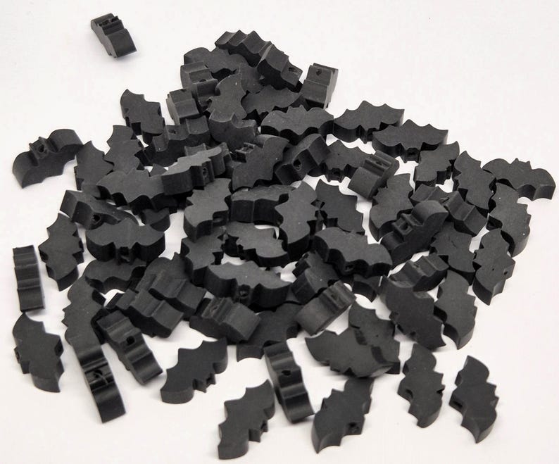 May include: A pile of small, black bat-shaped craft pieces scattered on a white surface. The bats have a simple, stylized design, with cut-out wings and a small body. These pieces are likely used for crafting or decoration.