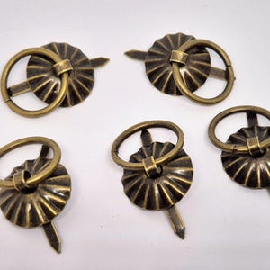 May include: Five antique gold-toned cabinet door pulls with a ring handle and a decorative fluted design. Each pull has a screw for installation.