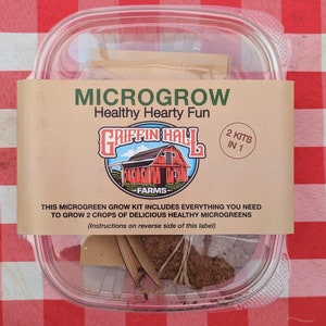 Pode incluir: Um recipiente de plástico transparente com dois kits de microgreens. O recipiente tem um rótulo castanho com o texto "MICROGROW Healthy Hearty Fun" e um logotipo de celeiro vermelho com o texto "GRIFFIN HALL FARMS". O rótulo também diz "2 KITS IN 1".