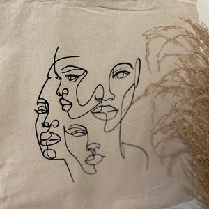 May include: A white canvas tote bag with a black line drawing of three overlapping faces. The faces are stylized and abstract.