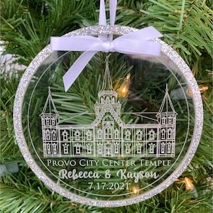 May include: A clear, round Christmas ornament with a silver glitter border. The ornament features a line drawing of the Provo City Center Temple with the text "Provo City Center Temple" and "Rebecca & Kayson 7.17.2021" below the drawing.