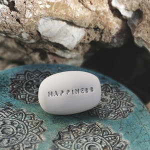 Puede incluir: Una piedra lisa y blanca con la palabra "HAPPINESS" grabada. Descansa sobre una superficie turquesa con un patrón de mandala. El fondo incluye una formación rocosa.