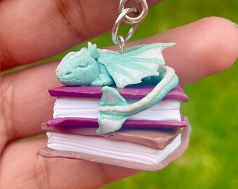 May include: A teal blue dragon charm with white wings sleeping on a stack of purple and white books. The charm is attached to a silver key chain.
