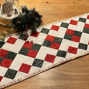 May include: A red and green quilted table runner with a diamond pattern. The runner has a white background with red and green squares. The runner has a fringed edge.