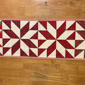 May include: A red and white quilted table runner with a starburst pattern. The runner has a red and white striped border.