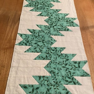 May include: A white table runner with a green leaf pattern. The pattern is made up of repeating chevron shapes. The runner is approximately 14 inches wide and 72 inches long.