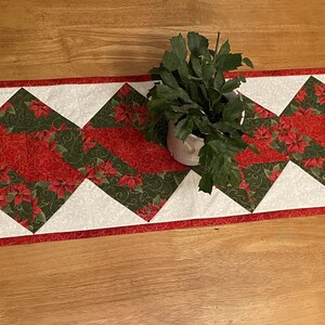 May include: A red and green Christmas table runner with a diamond pattern. The runner is made of fabric and has a white background with red and green poinsettia designs.