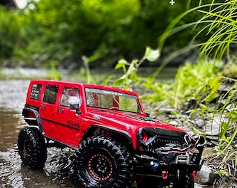 Può includere: Un modello di Jeep rossa con ruote nere e un paraurti anteriore nero è mostrato in un ruscello. La Jeep ha una griglia nera e un cofano nero. Lo sfondo è sfocato con fogliame verde.
