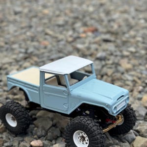 Könnte beinhalten: Ein hellblauer Spielzeug-LKW mit weißem Dach und schwarzen Reifen. Der LKW steht auf einem Bett aus kleinen Steinen.