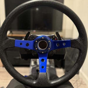 Könnte beinhalten: Ein schwarzes Rennsportlenkrad mit blauem Rand und Schaltwippen. Das Lenkrad ist an einem Rennsportsimulator befestigt.