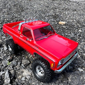 May include: A red toy truck with a black stripe and a silver roof rack with lights. The truck is sitting on a bed of gravel.