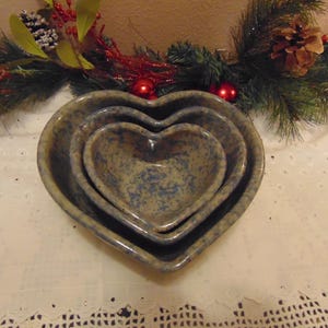 May include: A set of three heart-shaped nesting bowls in speckled blue and tan ceramic. The bowls are arranged in descending size, with a Christmas garland in the background.