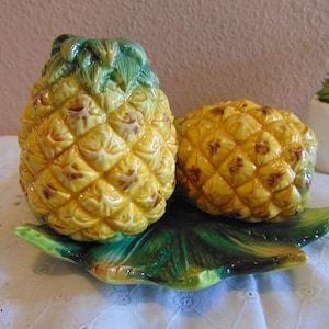 May include: A ceramic salt and pepper shaker set shaped like pineapples, featuring a yellow and brown textured fruit body with green leaves. The two pineapple shakers rest on a matching green ceramic leaf-shaped base with a glossy finish.