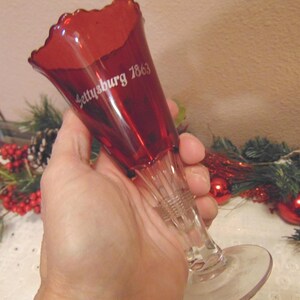 May include: A ruby red glass goblet with a flared rim and a clear stem. The goblet has the inscription "Gettysburg 1863" in white script. The goblet is held in a hand, with holiday decorations in the background.