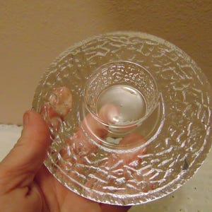 May include: Clear glass candle holder with a textured, circular design. The candle holder has a small, round center for the candle and a larger, textured base. The glass is transparent, allowing light to pass through.