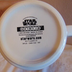 May include: The white underside of a ceramic mug featuring black printed text that reads STAR WARS, Galerie, 1-859-538-8500, MADE IN CHINA, Manufactured and distributed under license by GALERIE, starwars.com, &copy; & &trade; Lucasfilm Ltd., 34123 1349-1161 197.