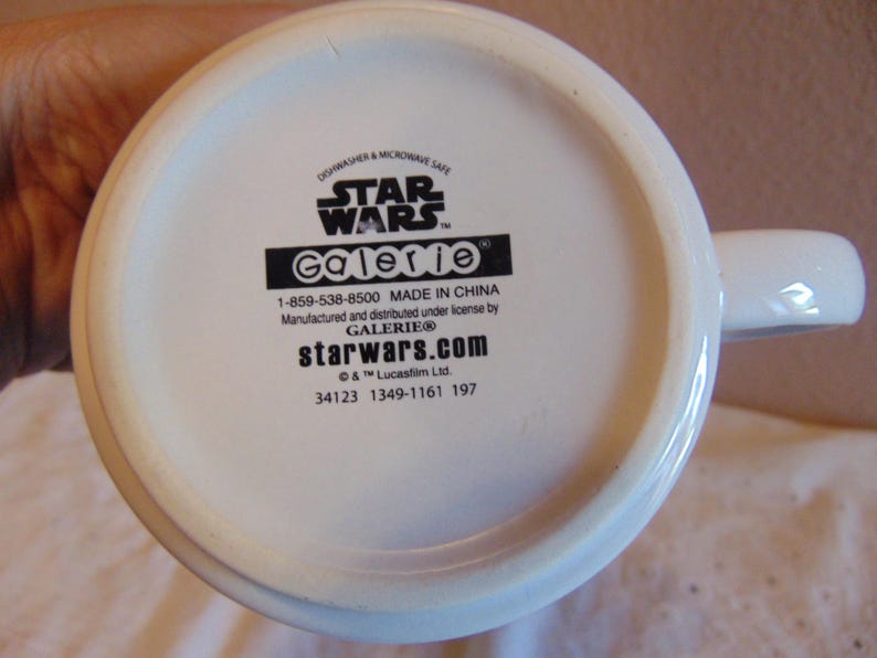 May include: The white underside of a ceramic mug featuring black printed text that reads STAR WARS, Galerie, 1-859-538-8500, MADE IN CHINA, Manufactured and distributed under license by GALERIE, starwars.com, &copy; & &trade; Lucasfilm Ltd., 34123 1349-1161 197.