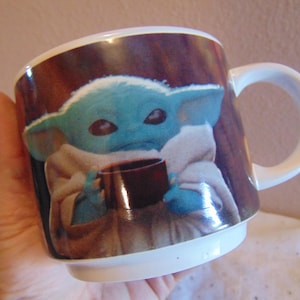 May include: A white ceramic mug featuring a printed graphic of a small, blue-skinned creature with large ears and dark eyes, wrapped in a beige robe and holding a small brown cup. The mug has a smooth, glossy finish and a curved handle on the side.