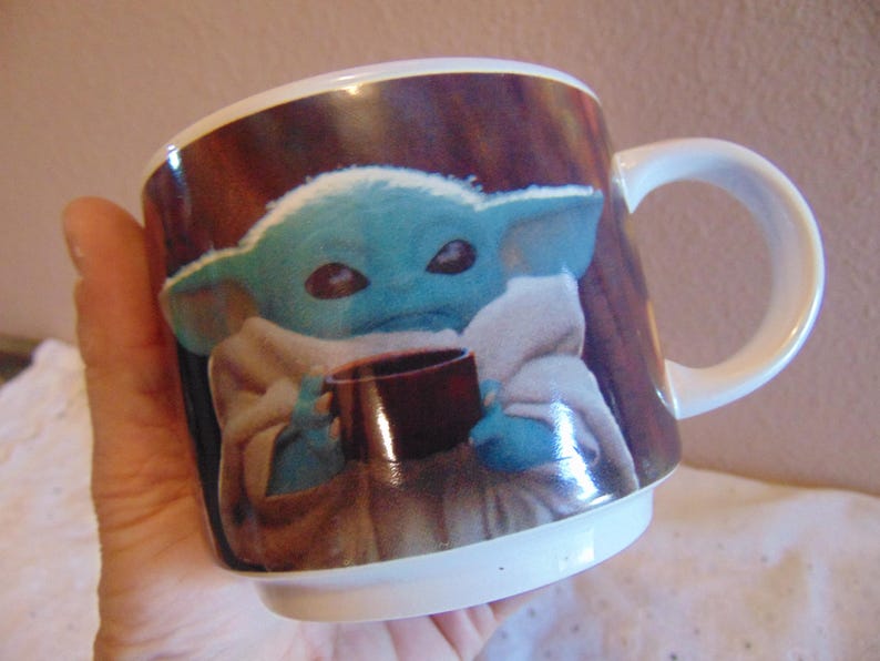 May include: A white ceramic mug featuring a printed graphic of a small, blue-skinned creature with large ears and dark eyes, wrapped in a beige robe and holding a small brown cup. The mug has a smooth, glossy finish and a curved handle on the side.