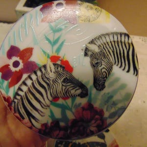 Pu&ograve; includere: Un oggetto decorativo rotondo e bianco con un disegno di due zebre a strisce bianche e nere. Il design include fiori rossi, viola e bianchi, insieme a foglie verdi. L'oggetto ha una finitura lucida e un tema floreale e animale.