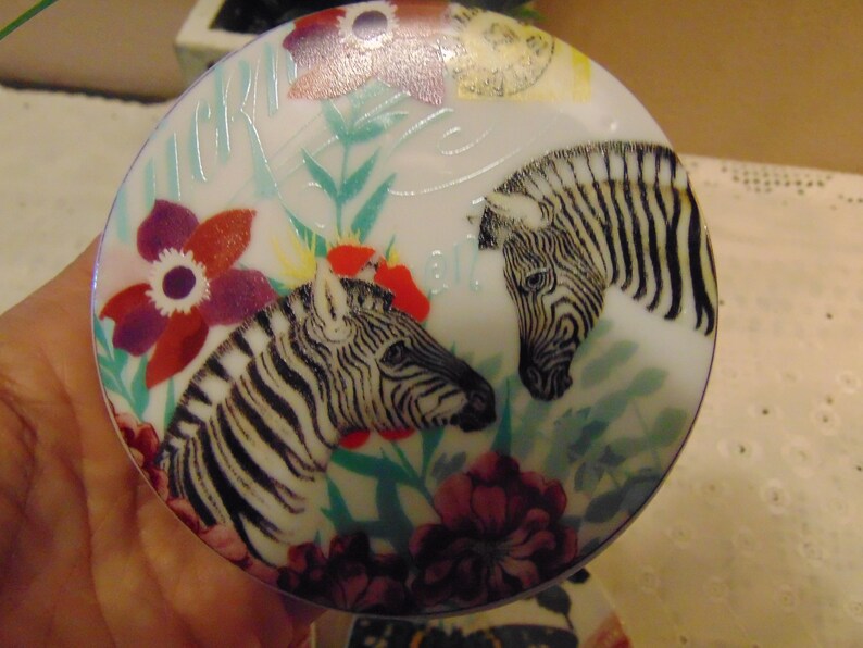 Pu&ograve; includere: Un oggetto decorativo rotondo e bianco con un disegno di due zebre a strisce bianche e nere. Il design include fiori rossi, viola e bianchi, insieme a foglie verdi. L'oggetto ha una finitura lucida e un tema floreale e animale.