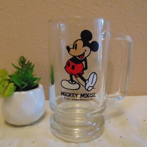 May include: A clear glass mug featuring a graphic of Mickey Mouse in black, red, and white. The text MICKEY MOUSE and WALT DISNEY PRODUCTIONS is printed in black below the character. The mug has a sturdy handle and a thick, raised base design.