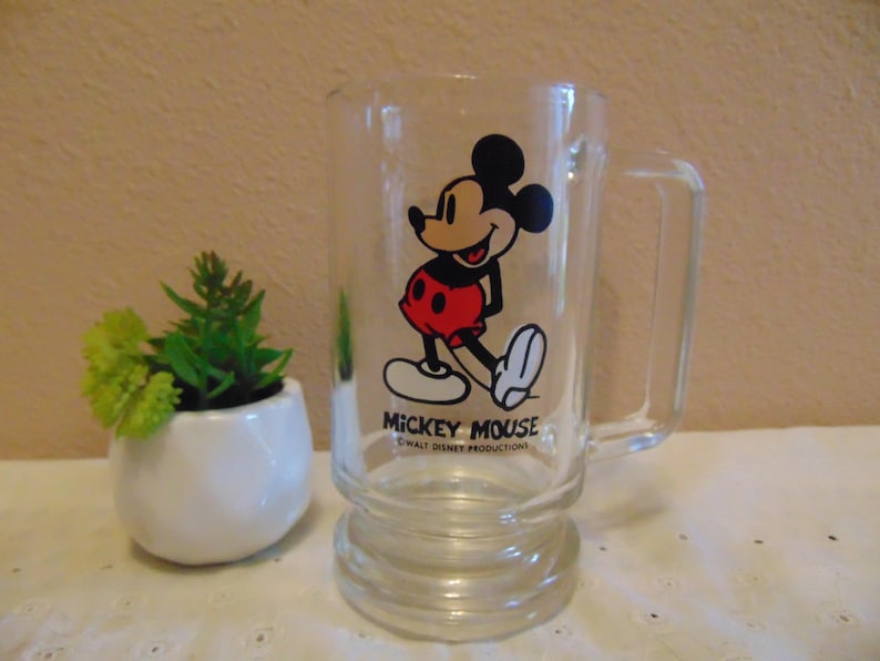 May include: A clear glass mug featuring a graphic of Mickey Mouse in black, red, and white. The text MICKEY MOUSE and WALT DISNEY PRODUCTIONS is printed in black below the character. The mug has a sturdy handle and a thick, raised base design.