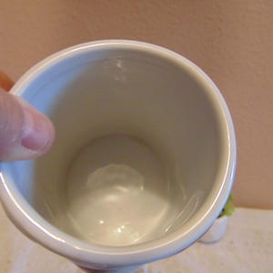 May include: A white ceramic mug with a smooth, glossy interior finish and a cylindrical shape. The mug features a simple, clean design with a rounded rim and a deep, hollow center, showcasing the plain white surface of the vessel's interior.