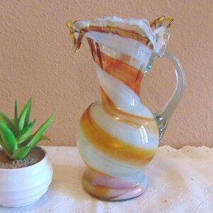 May include: A decorative glass pitcher with a handle, featuring a swirling pattern of white, orange, and yellow. The pitcher is next to a small white pot with a green succulent plant.