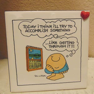 May include: A white ceramic tile with a cartoon illustration and text. The image features a character contemplating a calendar, with the text "TODAY I THINK I'LL TRY TO ACCOMPLISH SOMETHING...LIKE GETTING THROUGH IT!!!" A small red heart is attached to the top right corner.