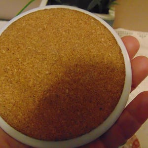 Pu&ograve; includere: Un sottobicchiere rotondo con base in sughero e bordo in ceramica bianca. La base in sughero &egrave; di colore marrone chiaro e ha una superficie strutturata. Il sottobicchiere &egrave; tenuto in mano.