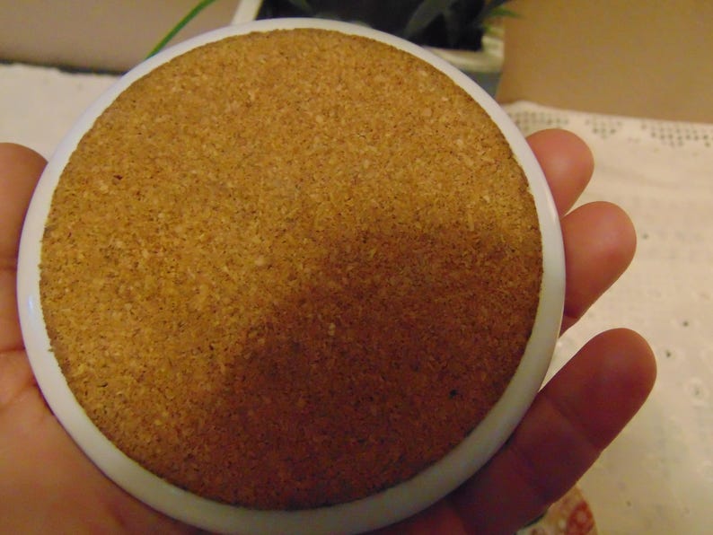 Pu&ograve; includere: Un sottobicchiere rotondo con base in sughero e bordo in ceramica bianca. La base in sughero &egrave; di colore marrone chiaro e ha una superficie strutturata. Il sottobicchiere &egrave; tenuto in mano.