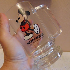 May include: A clear glass beer mug featuring a graphic of Mickey Mouse in red shorts and yellow shoes. The text MICKEY MOUSE and WALT DISNEY PRODUCTIONS is printed in black below the character. The mug has a sturdy handle and a thick base.