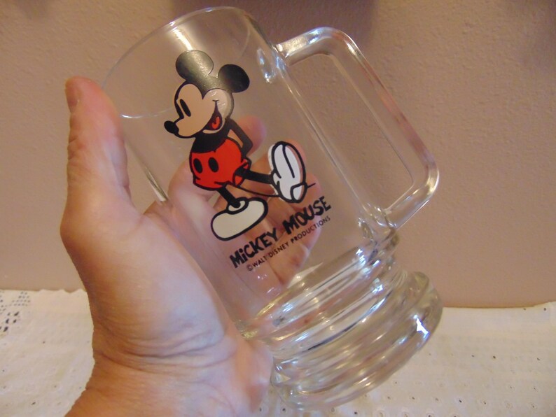 May include: A clear glass beer mug featuring a graphic of Mickey Mouse in red shorts and yellow shoes. The text MICKEY MOUSE and WALT DISNEY PRODUCTIONS is printed in black below the character. The mug has a sturdy handle and a thick base.