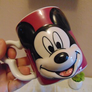 May include: A ceramic mug featuring a raised, three-dimensional relief of Mickey Mouse's face on a deep red background. The mug has a white interior and a white handle shaped with a curved design. The character has black ears, a white face.