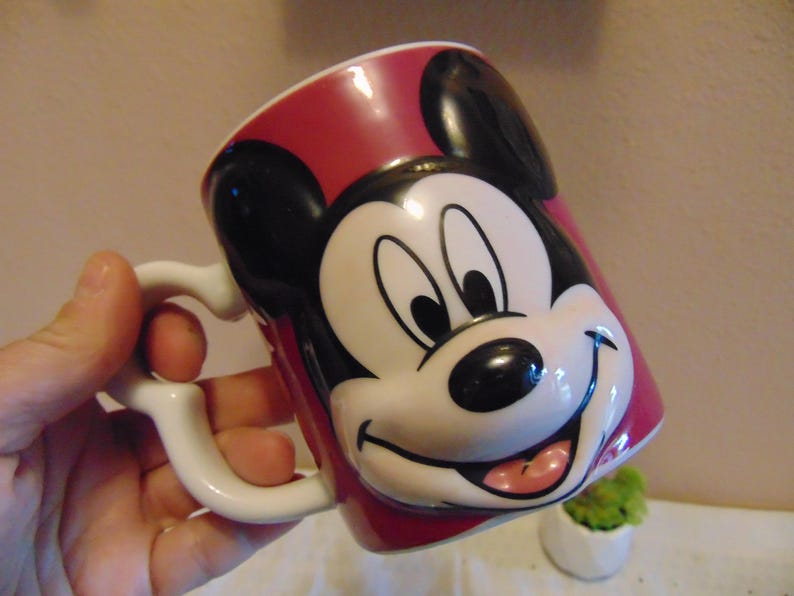 May include: A ceramic mug featuring a raised, three-dimensional relief of Mickey Mouse's face on a deep red background. The mug has a white interior and a white handle shaped with a curved design. The character has black ears, a white face.