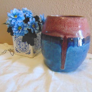 May include: A handmade ceramic vase with a two-tone glaze. The top is a reddish-brown, transitioning to a deep blue at the base. A small square ceramic pot with blue flowers is to the left of the vase.