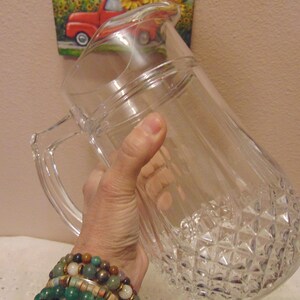 May include: A clear glass pitcher with a diamond-cut pattern on the bottom and a handle. The pitcher is held at an angle, showcasing its design. The pitcher is likely used for serving beverages.