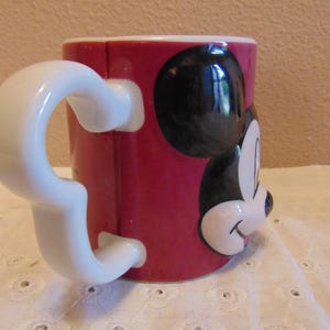 May include: A ceramic mug with a vibrant red exterior featuring a raised, three-dimensional relief of Mickey Mouse's face on the front. The mug includes a distinctively shaped white handle that mimics the silhouette of Mickey Mouse's ears.
