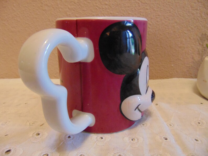 May include: A ceramic mug with a vibrant red exterior featuring a raised, three-dimensional relief of Mickey Mouse's face on the front. The mug includes a distinctively shaped white handle that mimics the silhouette of Mickey Mouse's ears.