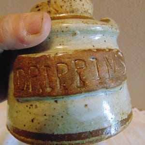 May include: A handmade ceramic jar with a cork stopper. The jar is beige with brown speckles and a brown band. The word "DRIPPINS" is embossed in a brown rectangular band around the jar.
