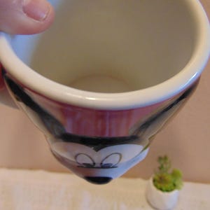 May include: A ceramic mug featuring a molded design of Mickey Mouse's face. The mug has a white interior and a handle on the side. The exterior displays the character's black ears, white face, and black nose, with large eyes positioned on the front.