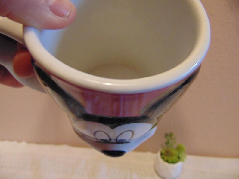 May include: A ceramic mug featuring a molded design of Mickey Mouse's face. The mug has a white interior and a handle on the side. The exterior displays the character's black ears, white face, and black nose, with large eyes positioned on the front.