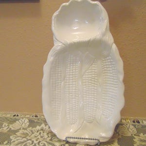 May include: A white ceramic serving dish shaped like a cluster of corn cobs. The piece features a textured surface with raised kernels and husks, including a rounded bowl-like section at the top and a larger, elongated tray section below.