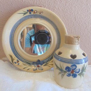 May include: A round, cream-colored mirror with blue and tan accents and floral details. Beside it is a matching ceramic bottle with heart-shaped cutouts and a cork stopper. Both items have blue floral designs.