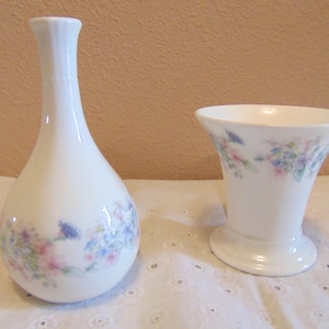 May include: Two white ceramic vases. One is a teardrop shape with a narrow neck, the other is a flared cylinder. Both vases are decorated with a floral pattern in pink, blue, and purple.