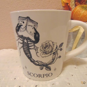 May include: A white ceramic mug featuring a black ink illustration of a scorpion holding an open book with star constellations, accompanied by a rose and the word SCORPIO printed in capital letters at the bottom of the front surface.